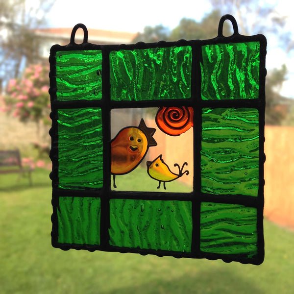 A BIRDS LOVE Hand Painted Glass Suncatcher created with green coloured glass framing the hand-painted clear glass featuring mama bird and her baby gazing at each other with a spiral sun in the background. The picture features two loops so it can be suspended in air or put on the wall, made by Southampton-based artist Lenka and LeKoky brand. Shot against a garden background to showcase its transparency.