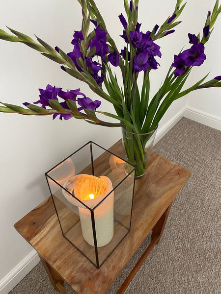 Create a cozy and inviting ambiance with this exquisite glass candle holder, handmade by a skilled stained glass artist in Lekoky, Southampton, Hampshire, England. Featuring a beautifully lit candle and a lovely bouquet of flowers, this piece is perfect for adding a touch of elegance to your home decor. The black patina finish on the soldering seams adds a unique touch to the lighting and creates a warm and inviting atmosphere.